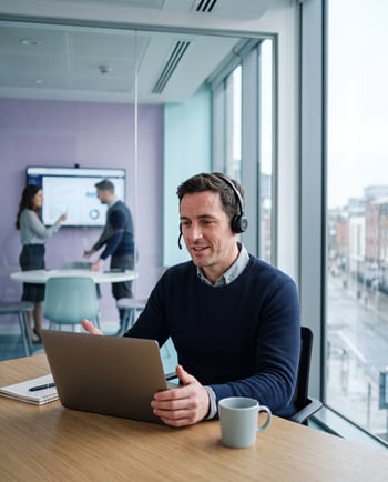 Team member on a video call in a hybrid office, with colleagues collaborating in the background
